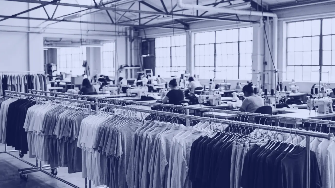 Rails of plain ready-to-wear garments in a factory with a sewing line in the background