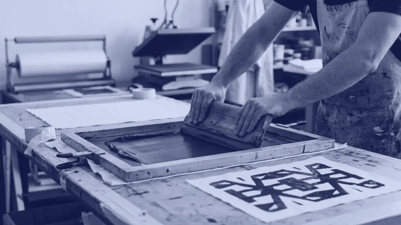 Screen printing table with a squeegee pulling blue ink through a frame onto white fabric beside a printed sample