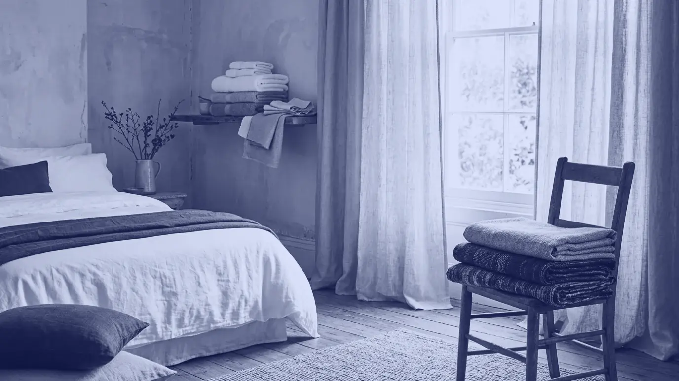 White bedding, folded towels on a shelf, linen curtains and folded upholstery fabric on a chair