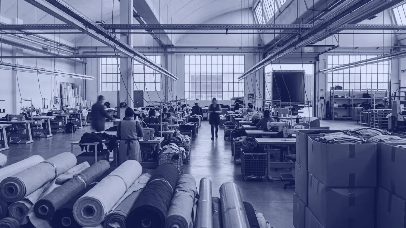 Wide garment factory hall in morning light with sewing stations, fabric rolls and cartons of finished garments