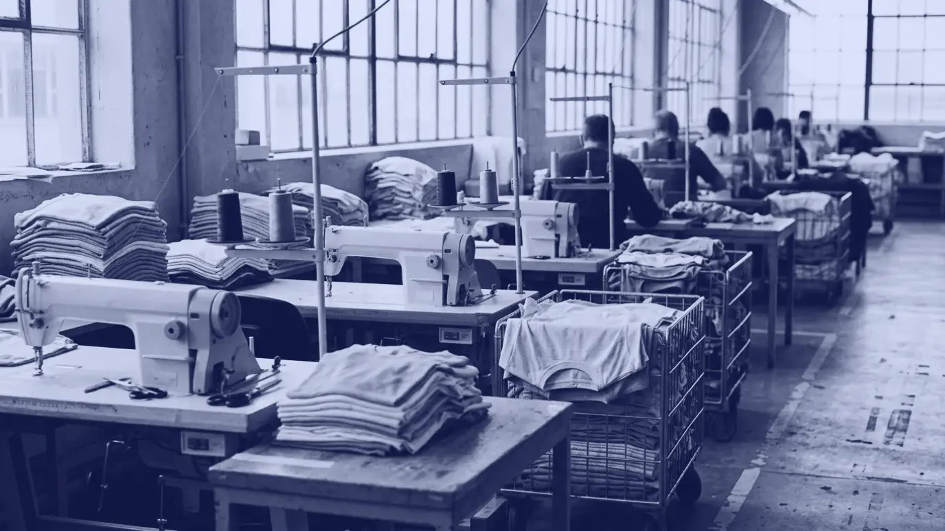 Sewing line in a clothing factory with machines, stacks of folded knit panels and operators seen from behind
