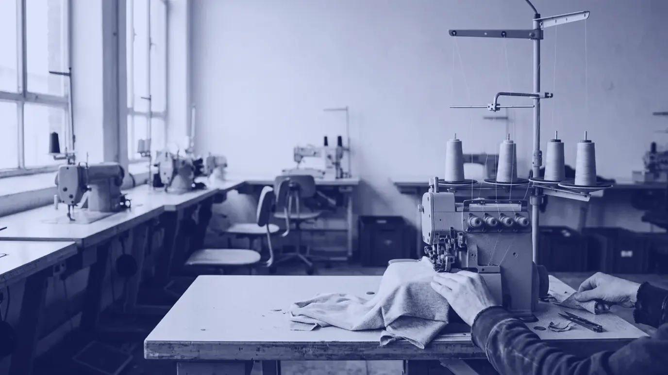 Hands guiding grey knit fabric through an overlock machine in a sewing workshop, with industrial machines lined up behind