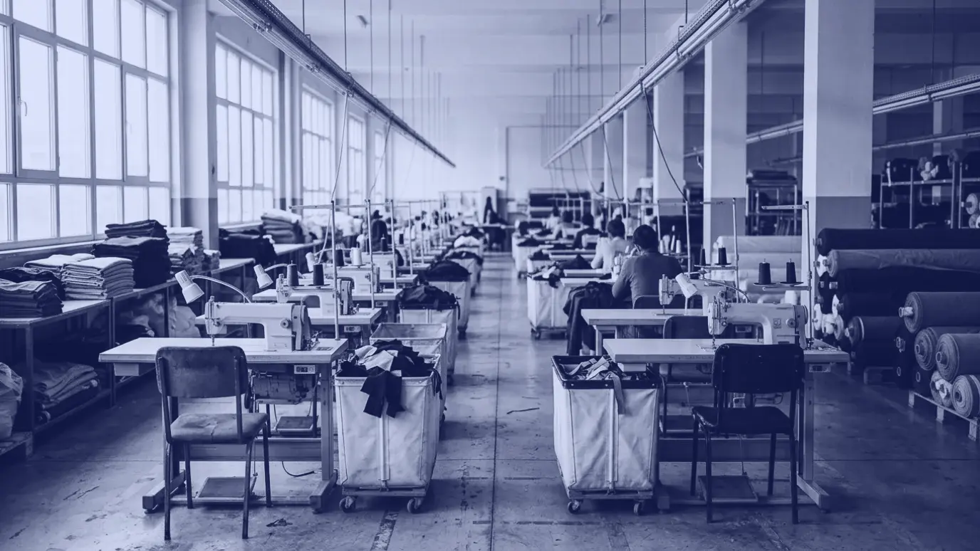 Konfeksiyon atölyesinde sıralı dikiş makineleri, bazı istasyonlar boş; yan duvarda kumaş topları ve katlı kumaşlar