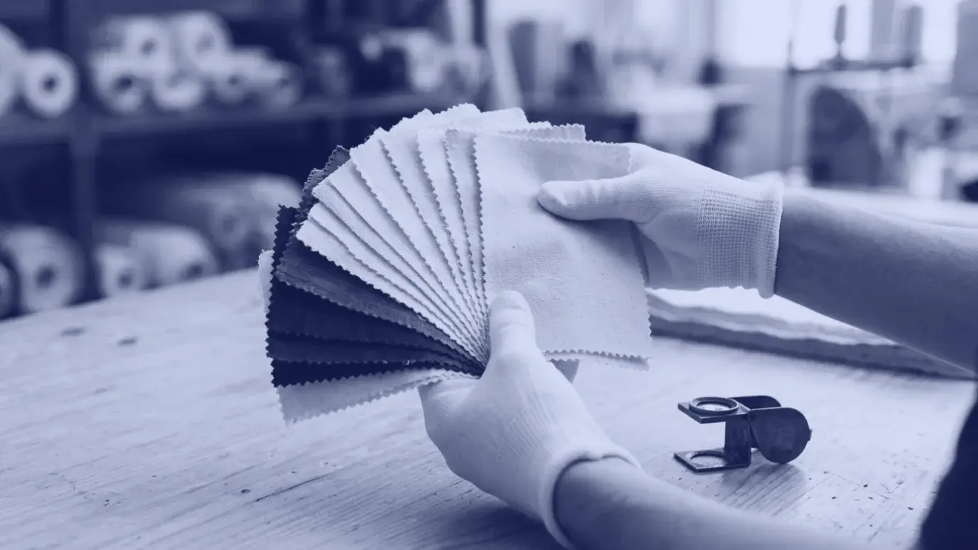 Gloved hands fanning out natural cotton and deep blue fabric swatches beside a brass magnifying loupe