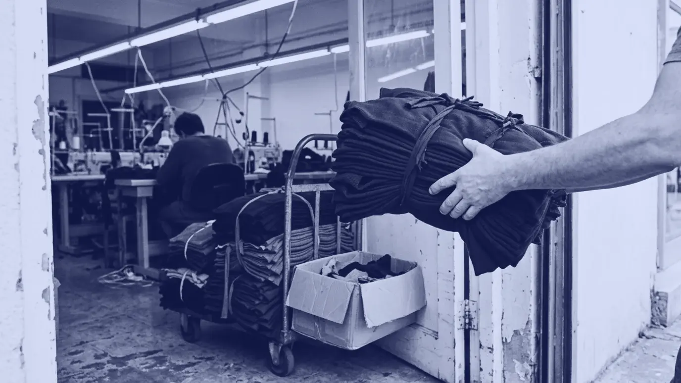 Atölye kapısında teslim edilen lacivert kesim parça demeti, yanında el arabası ve açık koli; arkada dikim makineleri