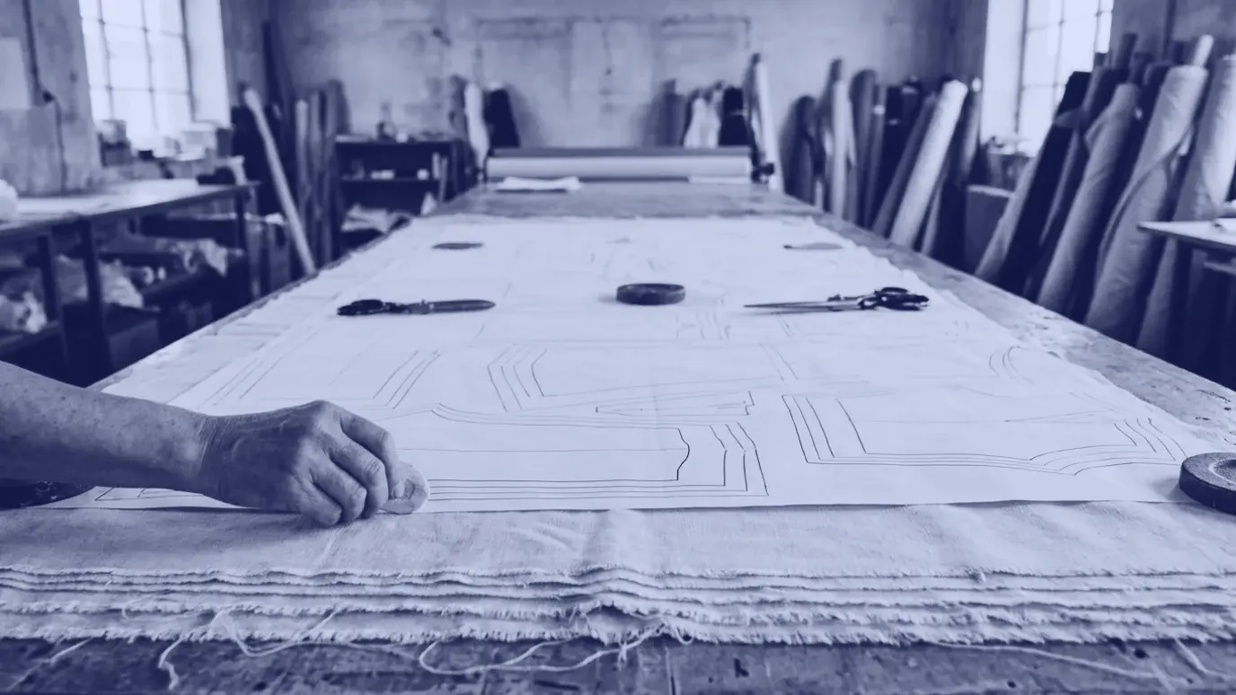 Long cutting table with spread fabric layers and a marker of tightly nested pattern pieces, a hand with chalk at the edge