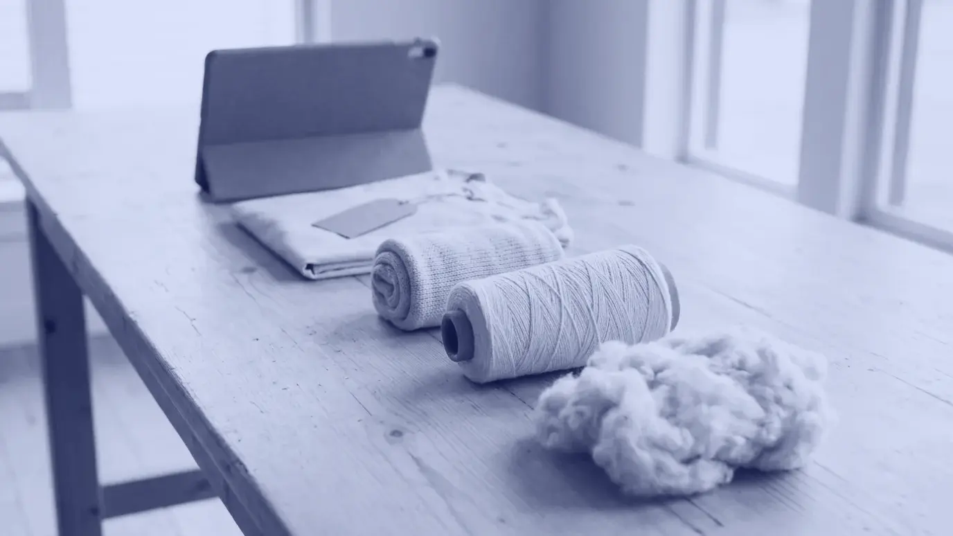 A tuft of raw cotton, a yarn cone, a rolled knit fabric and a folded tagged garment in a row on a table with a tablet behind