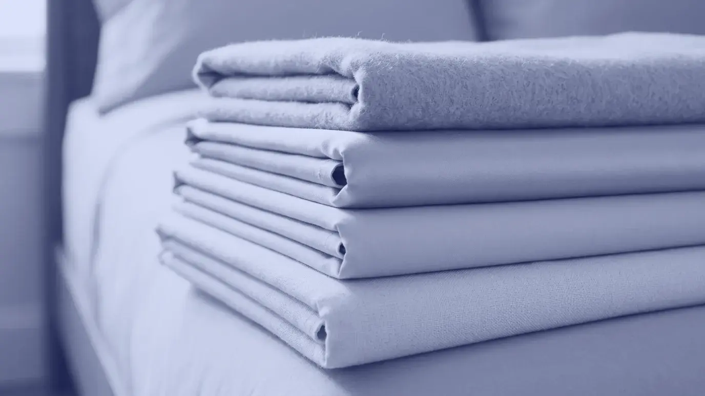 Stack of folded bed linen fabrics on a bed corner showing brushed, smooth and lustrous surfaces