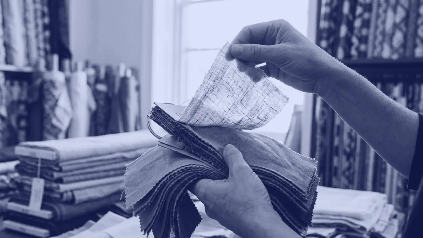 Hands comparing fabric swatches against window light in a showroom with fabric lengths blurred behind