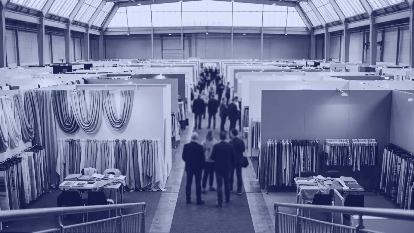 Visitors seen from behind walking between fabric booths in a large textile trade fair hall