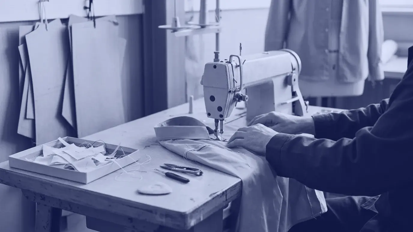 Numune atölyesinde, düz dikiş makinesinin altında yarım kalmış gri gömlek numunesini yönlendiren eller
