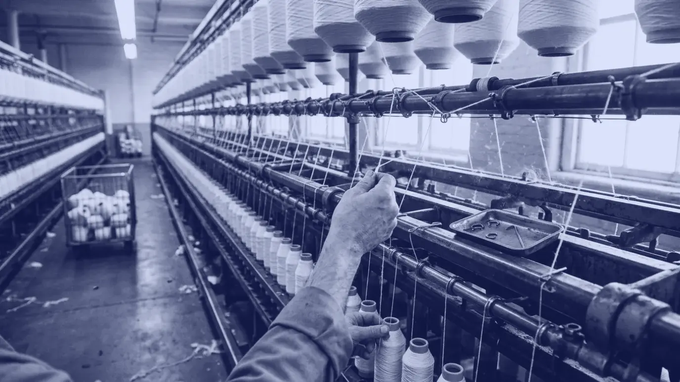 Ring iplik makinesinde kopan ipliğin ucunu tutan el; üstte fitil masuraları, altta sıralı iplik kopsları, havada pamuk lifi