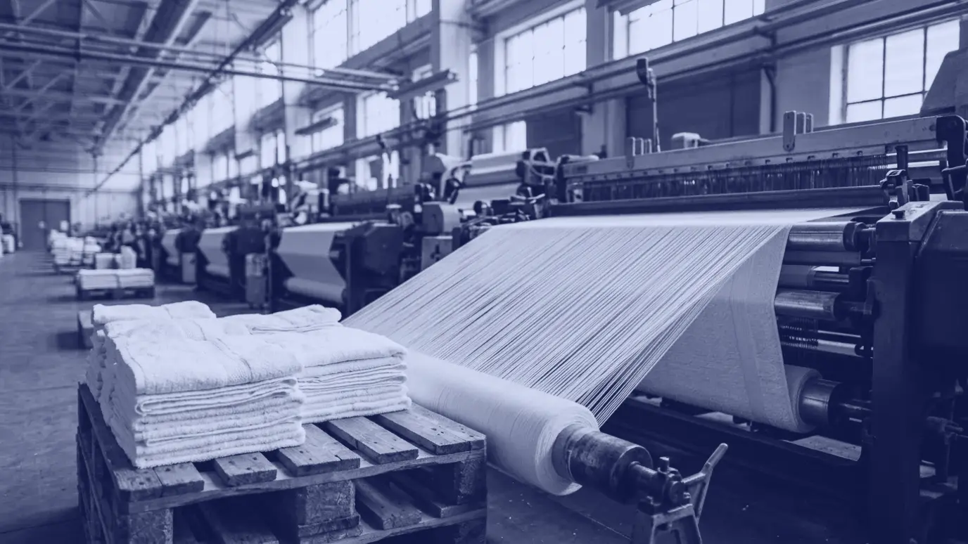 Dense white warp threads running into a terry towel loom, with stacks of folded white towels on a pallet in front