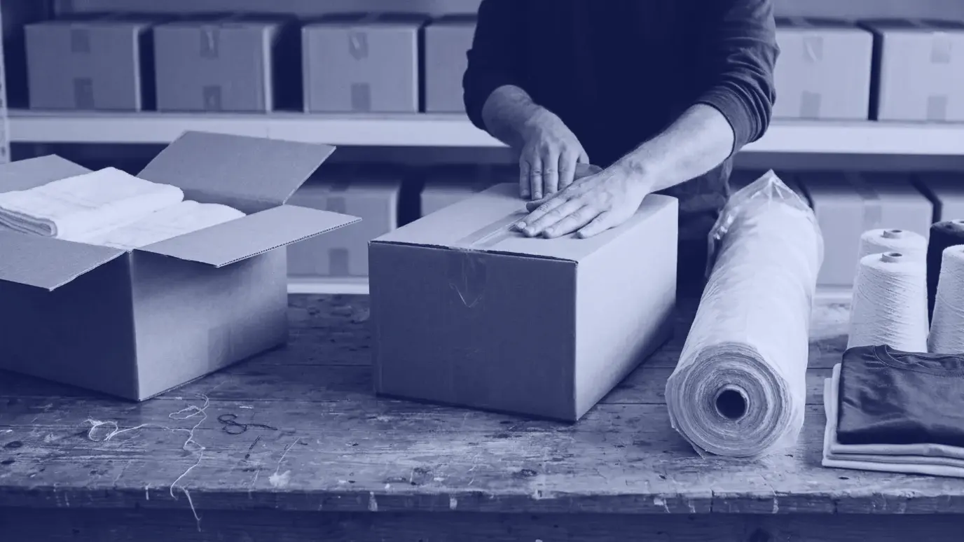 Depoda paketleme tezgâhında koliyi bantlayan eller; yanda havlu istifi, kumaş topu, iplik konileri ve katlı tişörtler
